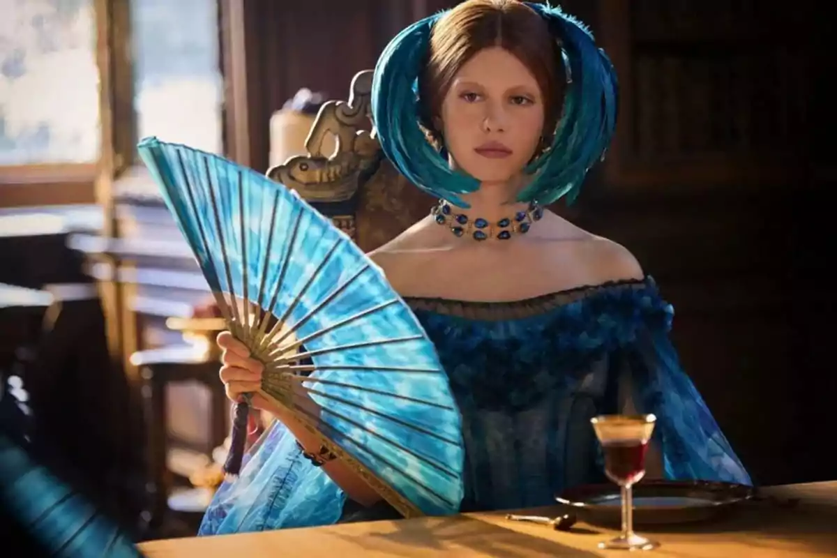 Mujer vestida con un elegante vestido azul y adornos de plumas en el cabello sostiene un abanico azul mientras está sentada en una mesa con una copa de vino Mujer vestida con un elegante vestido azul y adornos de plumas en el cabello sostiene un abanico azul mientras está sentada en una mesa con una copa de vino