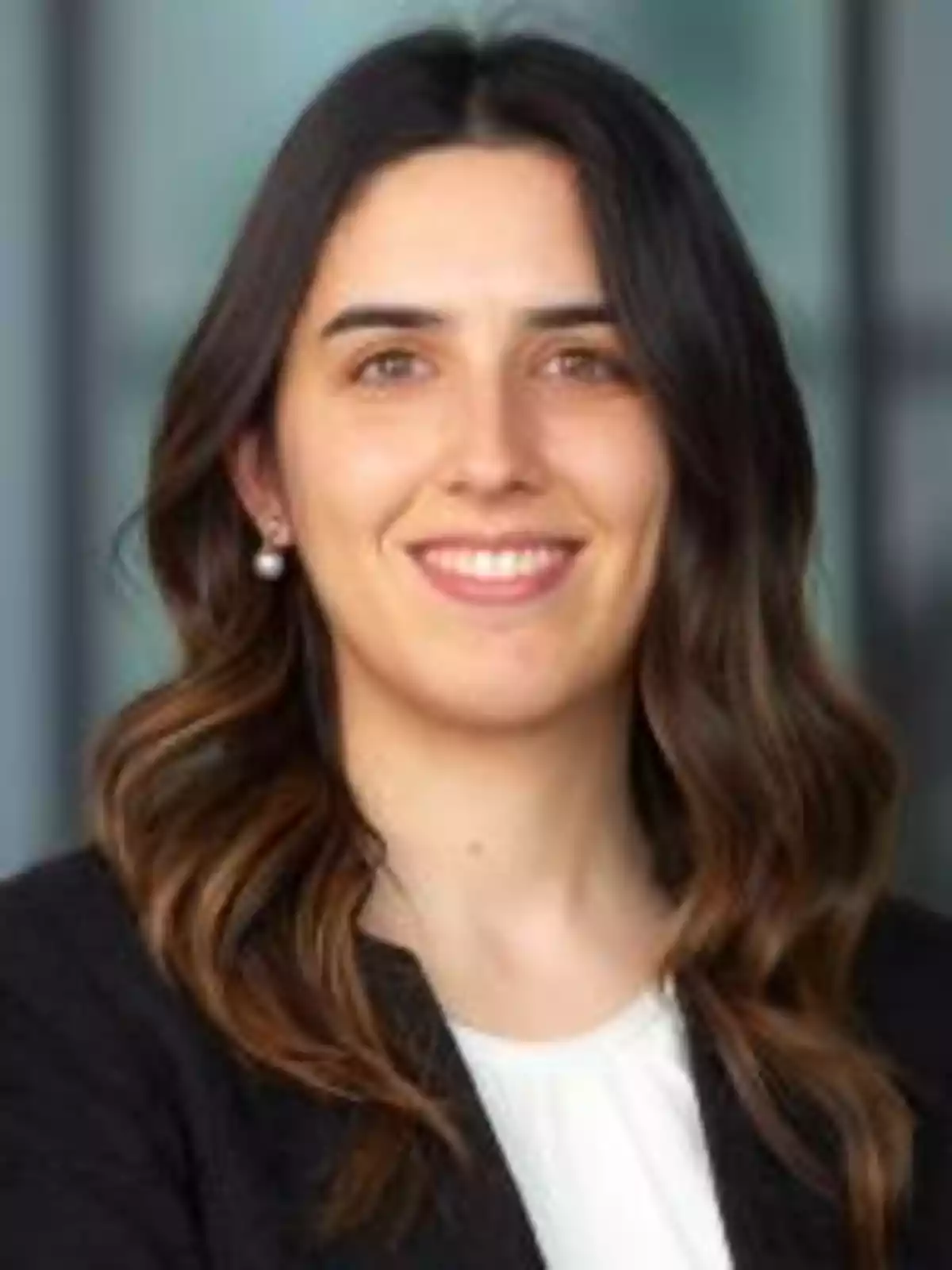 Mujer joven de cabello largo y ondulado con una chaqueta oscura y blusa blanca sonriendo frente a un fondo desenfocado Mujer joven de cabello largo y ondulado con una chaqueta oscura y blusa blanca sonriendo frente a un fondo desenfocado