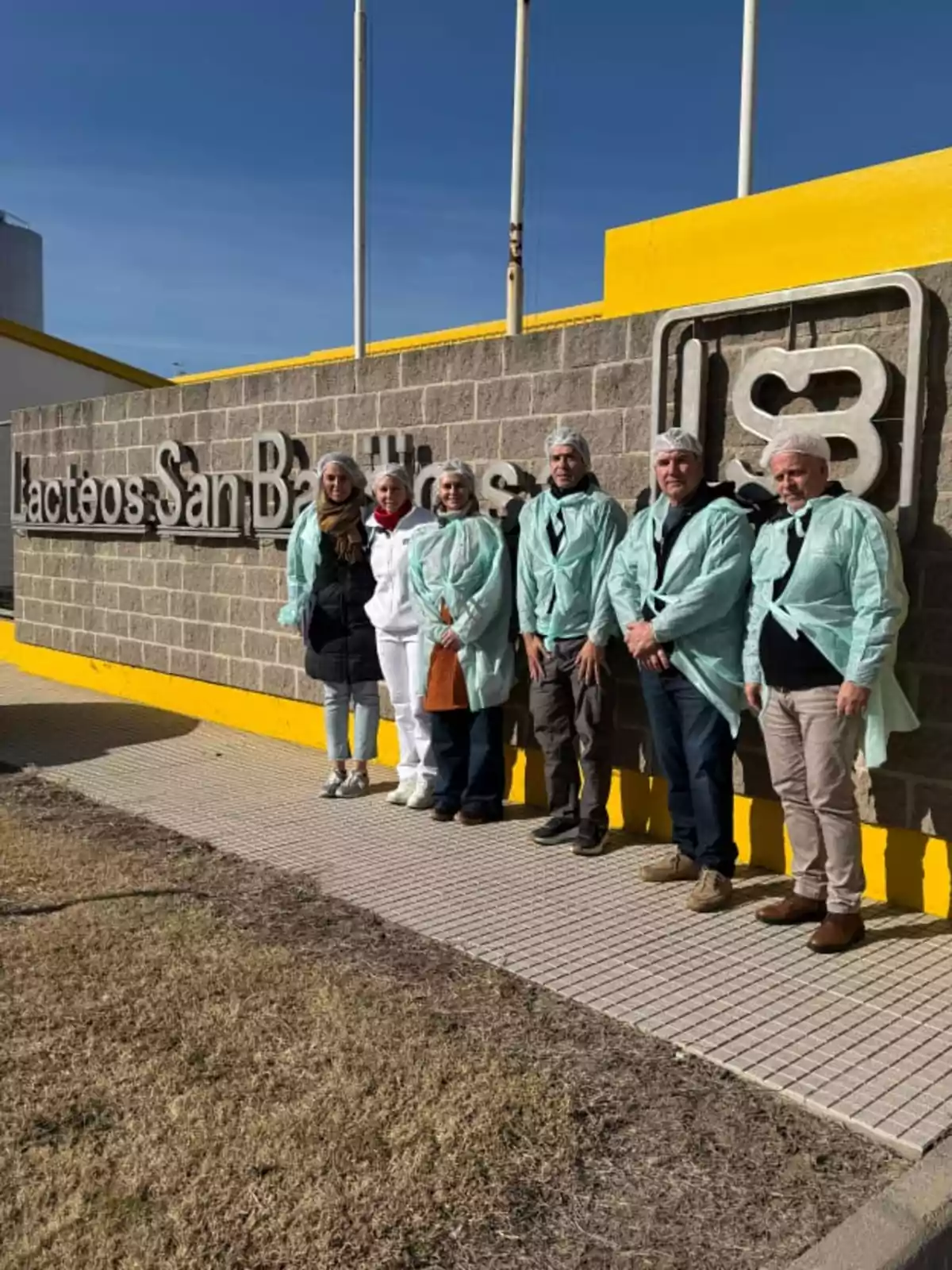 Grupo de personas con batas y cofias posando frente a un cartel de Lácteos San Basilio en un día soleado Grupo de personas con batas y cofias posando frente a un cartel de Lácteos San Basilio en un día soleado