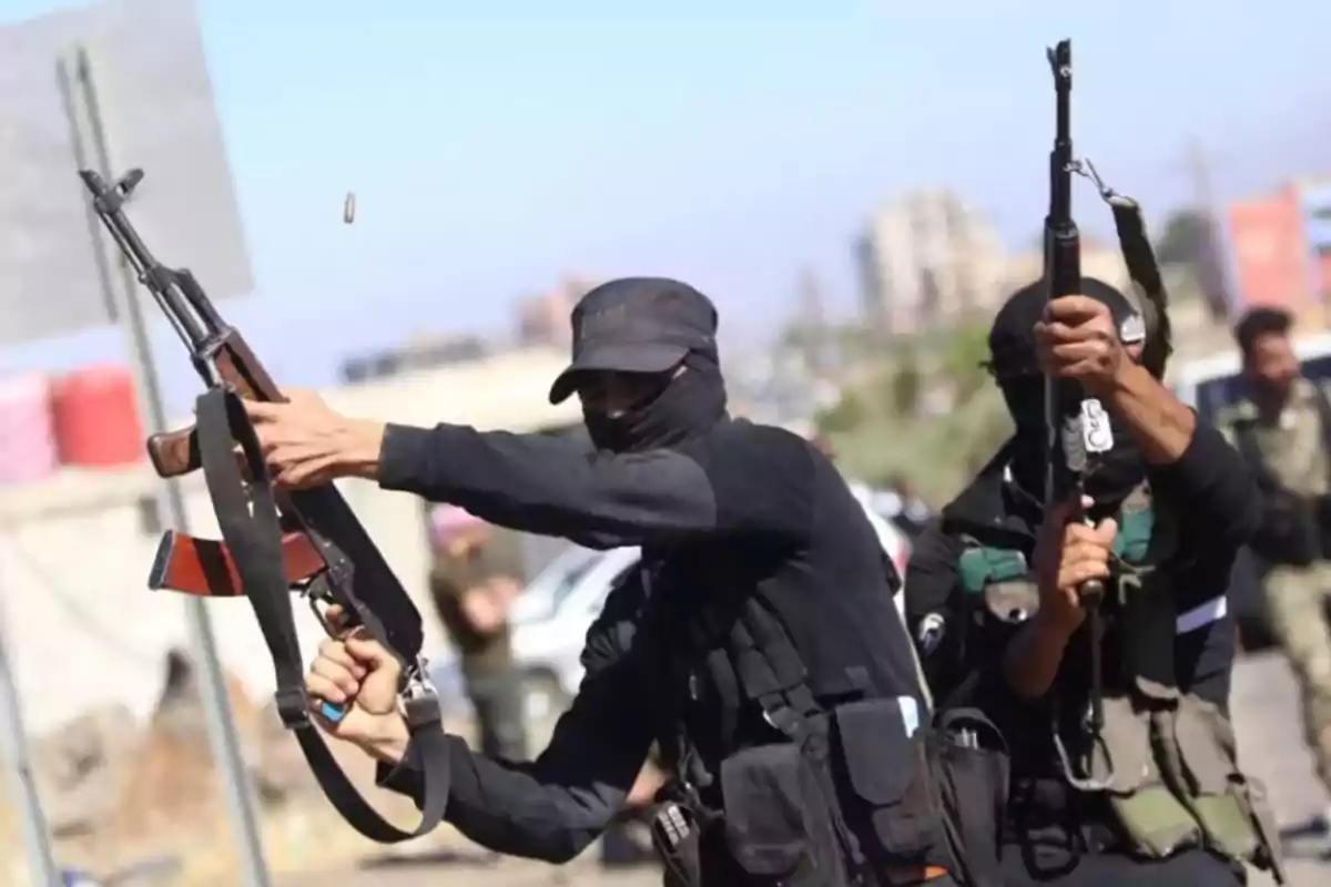 Dos personas encapuchadas vestidas de negro disparan rifles automáticos al aire en una calle durante el día Dos personas encapuchadas vestidas de negro disparan rifles automáticos al aire en una calle durante el día