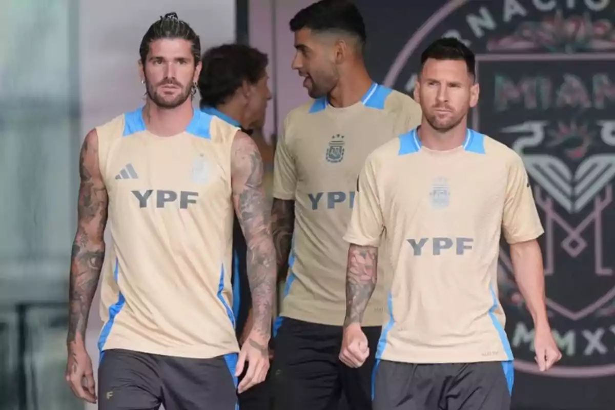 Tres futbolistas con uniforme de entrenamiento color beige y detalles celestes caminan juntos en un entorno deportivo Tres futbolistas con uniforme de entrenamiento color beige y detalles celestes caminan juntos en un entorno deportivo