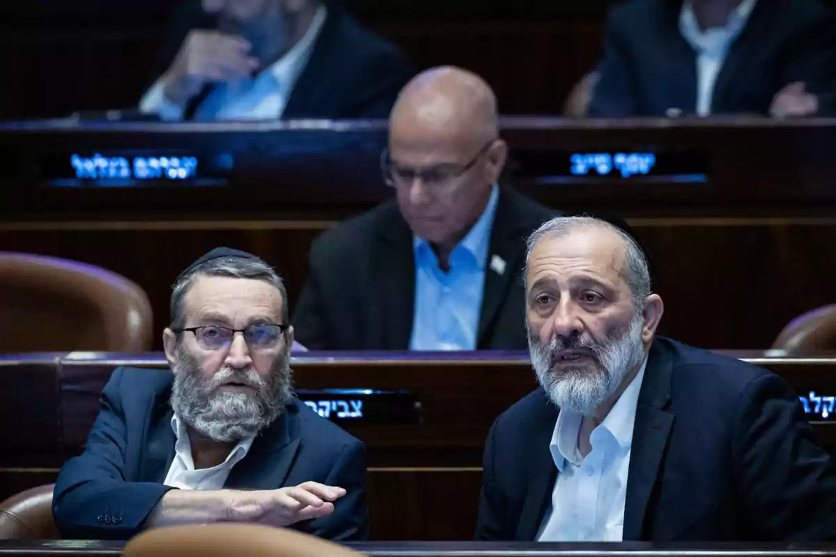 Two men with beards and kippahs are talking while seated in a parliamentary chamber, with other people in the background. Two men with beards and kippahs are talking while seated in a parliamentary chamber, with other people in the background.