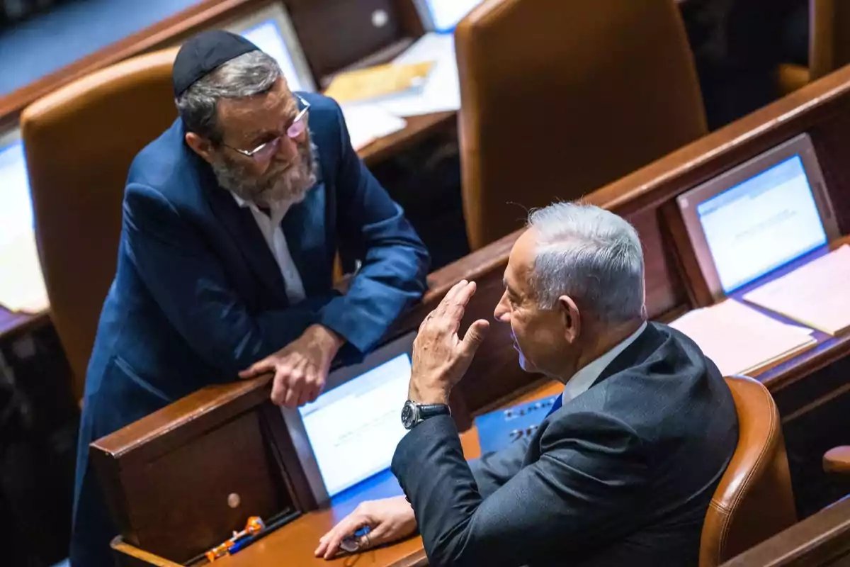 Two men are conversing in a parliamentary chamber, one standing with a kippah and a beard, the other sitting in a dark suit gesturing with his hand.