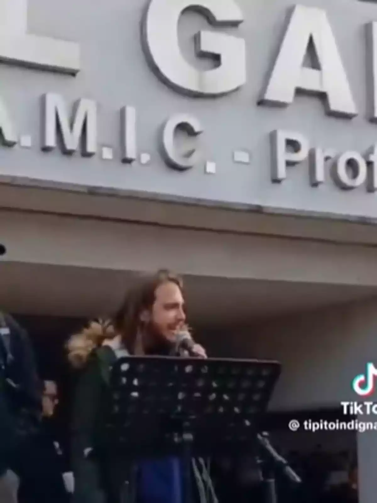 Persona hablando por micrófono frente a un atril en un evento al aire libre, con un letrero grande de fondo y varias personas alrededor. Persona hablando por micrófono frente a un atril en un evento al aire libre, con un letrero grande de fondo y varias personas alrededor.
