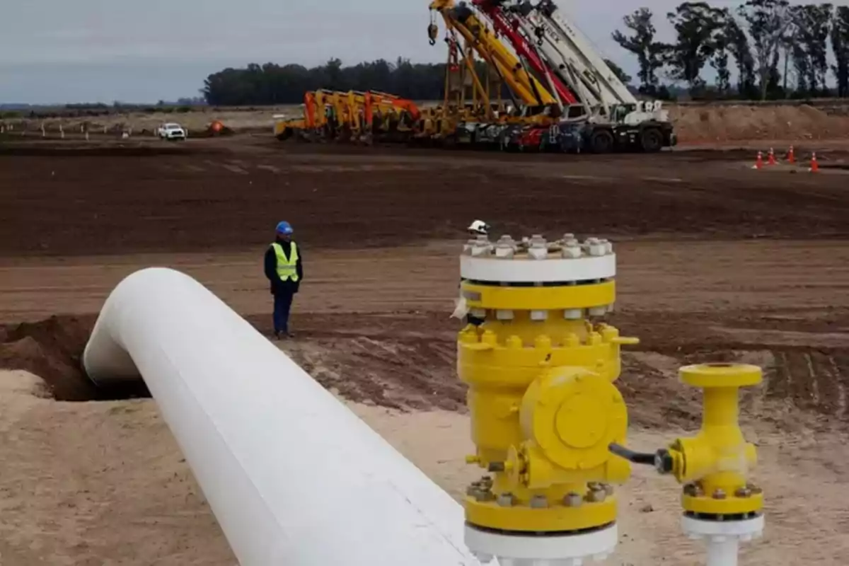 Trabajadores con casco y chaleco supervisan una tubería de gas blanca y amarilla en una zona de construcción con maquinaria pesada al fondo Trabajadores con casco y chaleco supervisan una tubería de gas blanca y amarilla en una zona de construcción con maquinaria pesada al fondo