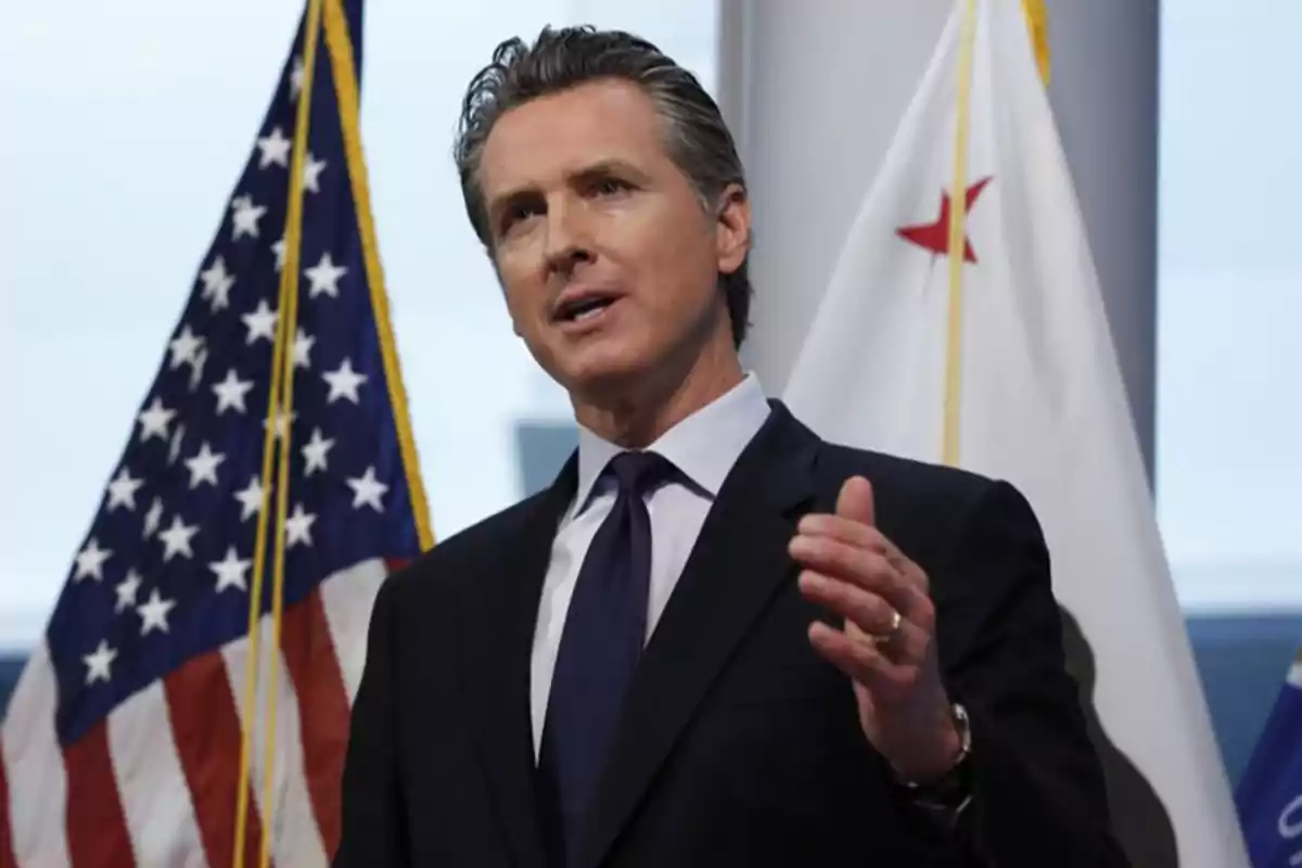 Hombre de traje hablando frente a micrófonos con banderas de Estados Unidos y California al fondo