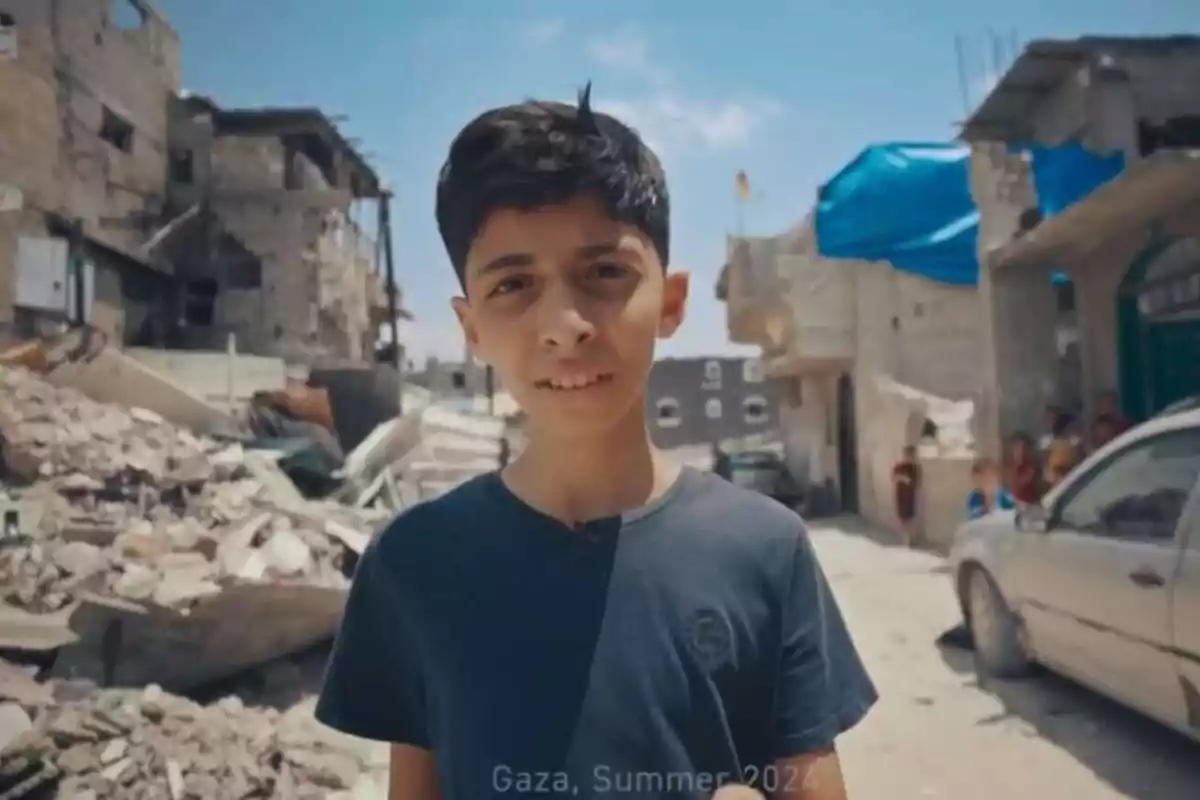 Un niño de cabello oscuro y camiseta azul está de pie en una calle rodeada de edificios dañados y escombros bajo un cielo despejado Un niño de cabello oscuro y camiseta azul está de pie en una calle rodeada de edificios dañados y escombros bajo un cielo despejado