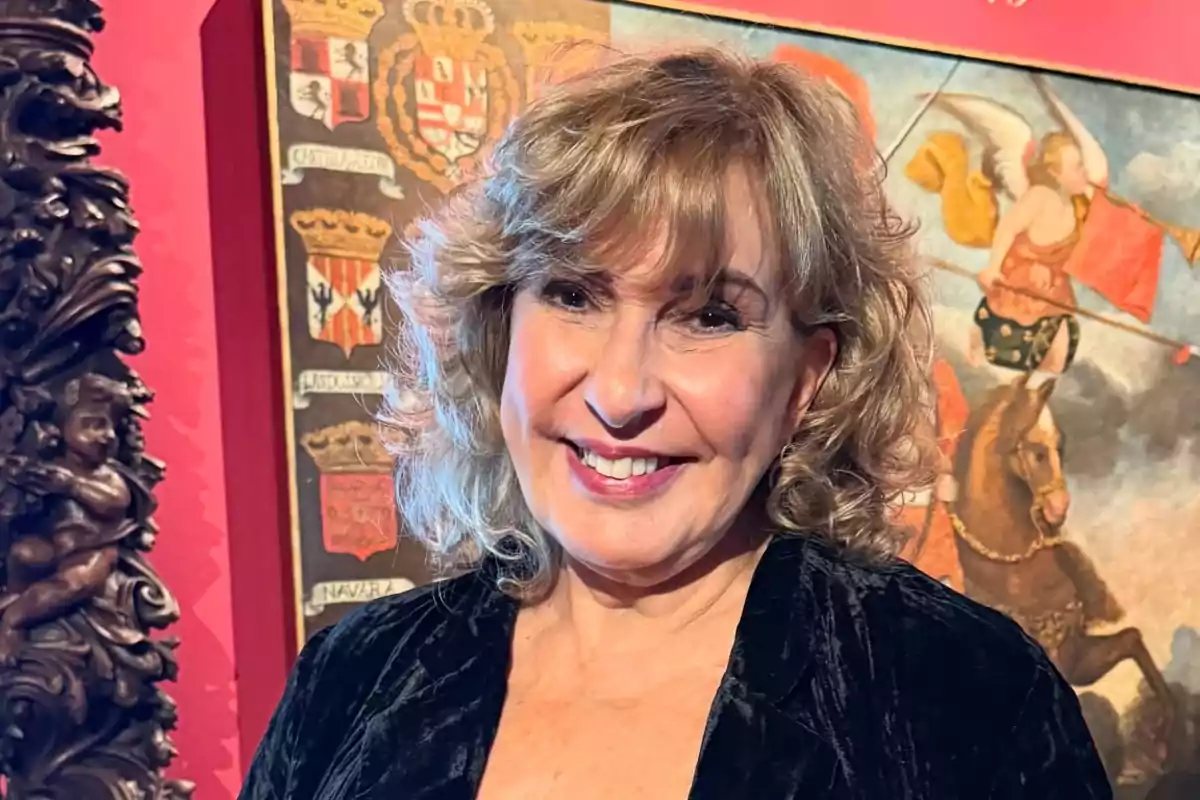 Blonde, curly-haired woman smiles in front of an old painting with shields and angelic figures in a museum setting.