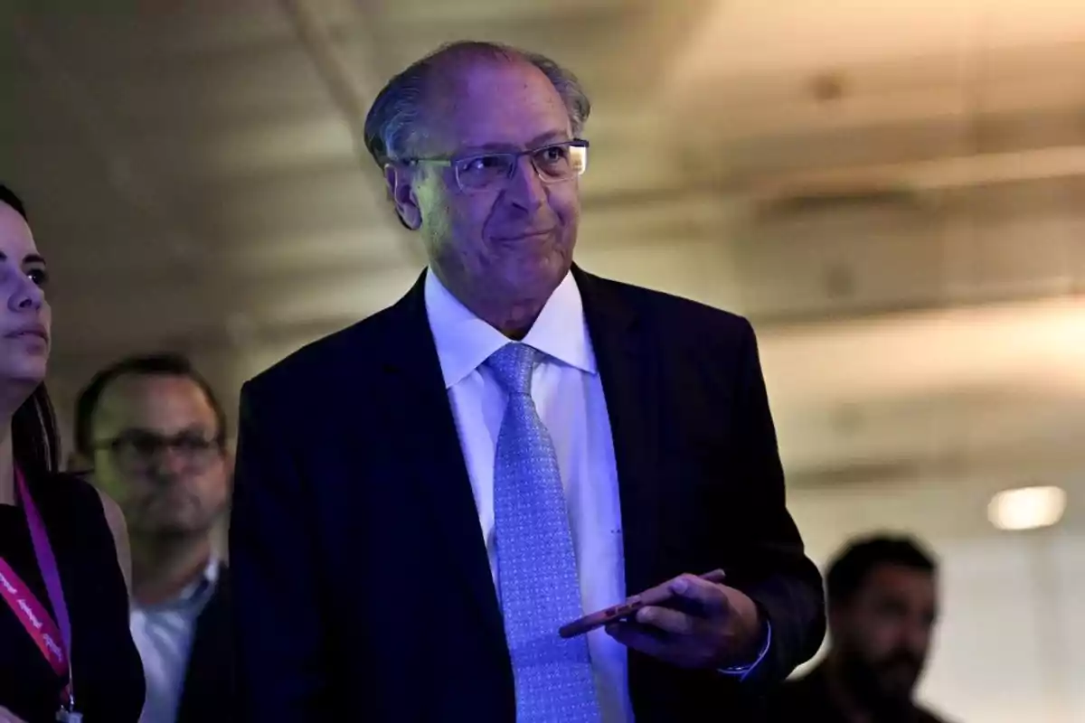 Older man with glasses and a dark suit holding a mobile phone in a dimly lit indoor setting with people in the background Older man with glasses and a dark suit holding a mobile phone in a dimly lit indoor setting with people in the background