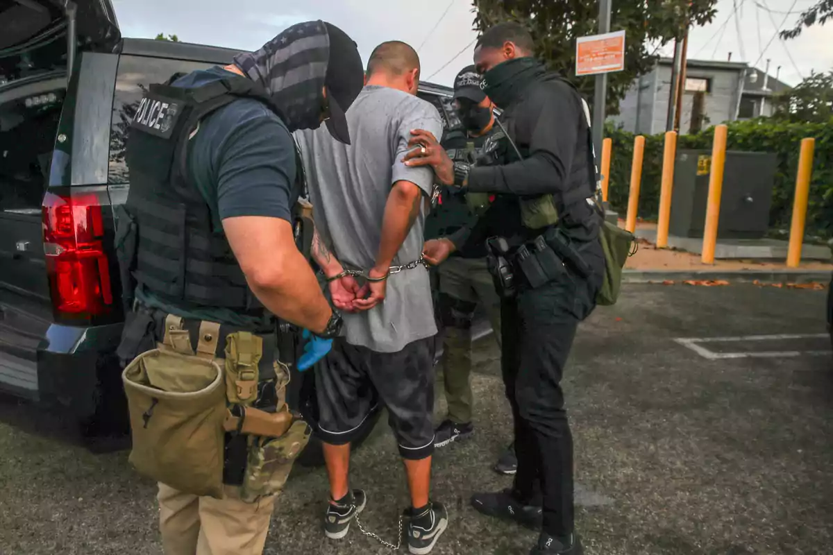 Three police officers arrest a handcuffed man next to a pickup truck in a parking lot. Three police officers arrest a handcuffed man next to a pickup truck in a parking lot.