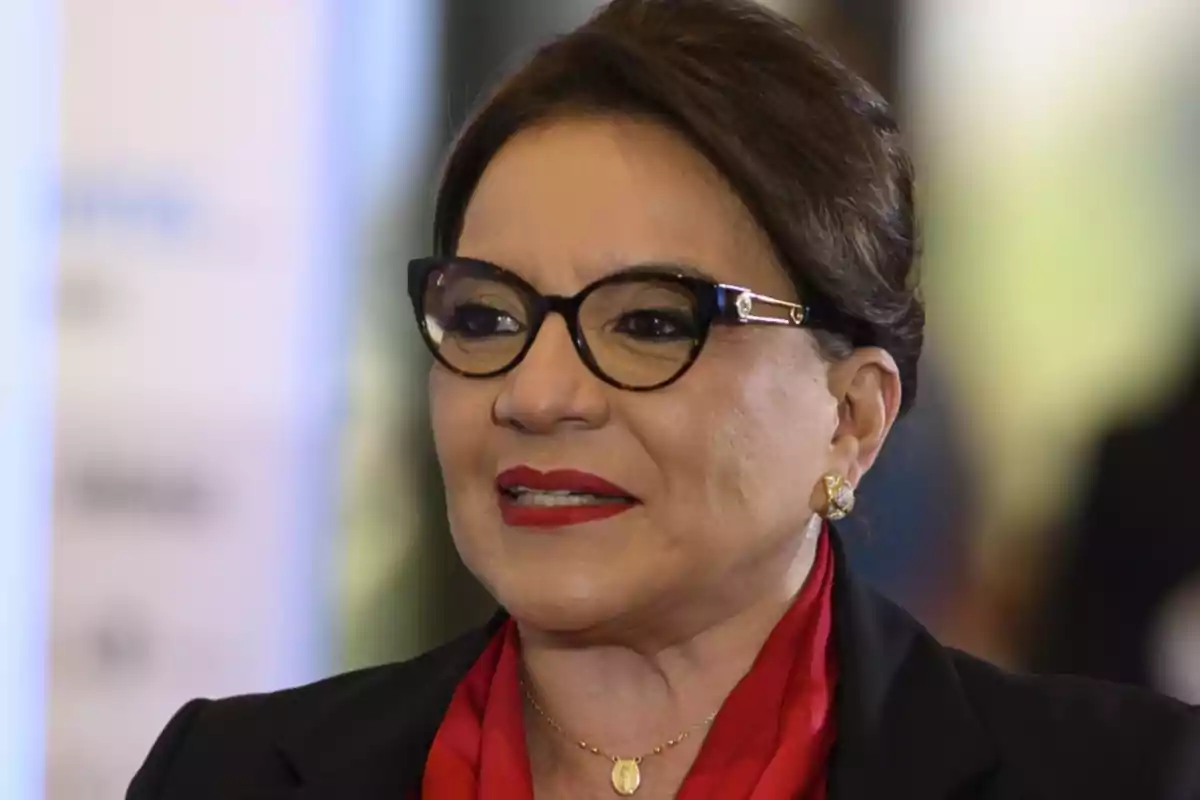Woman with her hair up, wearing dark sunglasses, red lipstick, and a red scarf, with a gold necklace and earrings, against a blurred background. Woman with her hair up, wearing dark sunglasses, red lipstick, and a red scarf, with a gold necklace and earrings, against a blurred background.