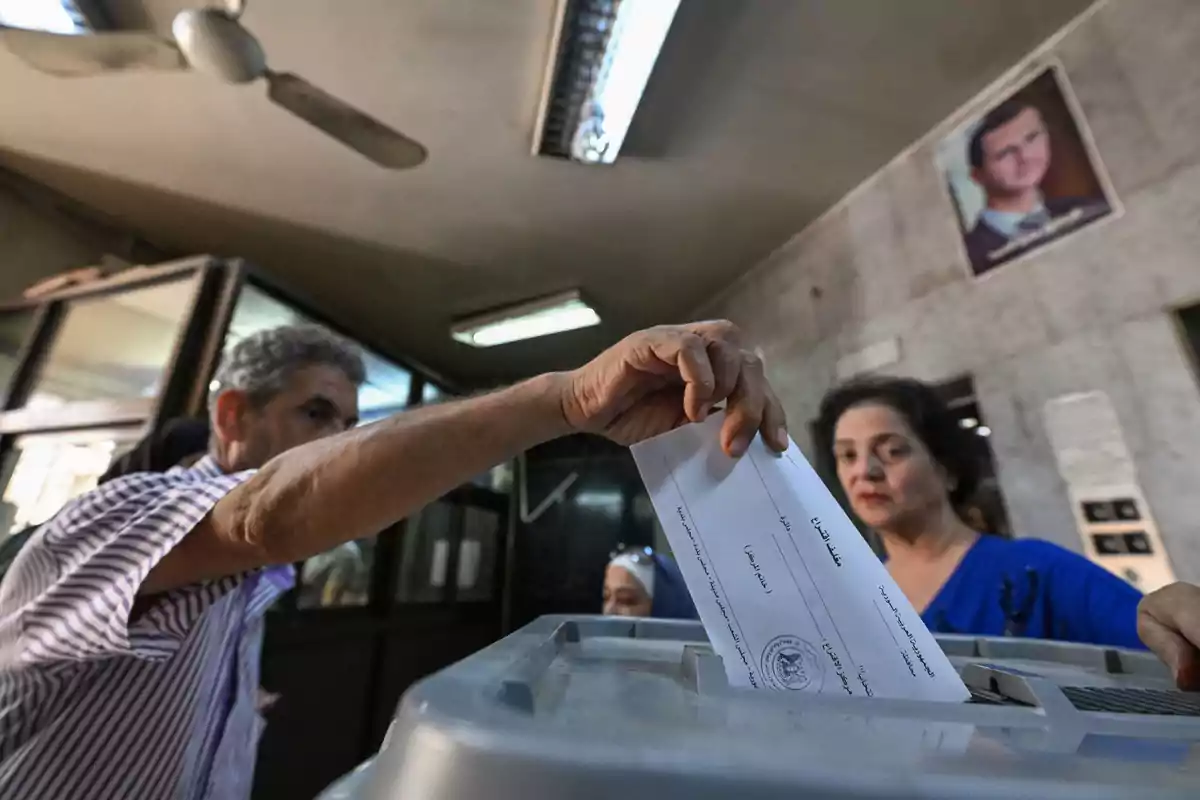 A man places a ballot into a voting box while a woman watches in an enclosed room. A man places a ballot into a voting box while a woman watches in an enclosed room.