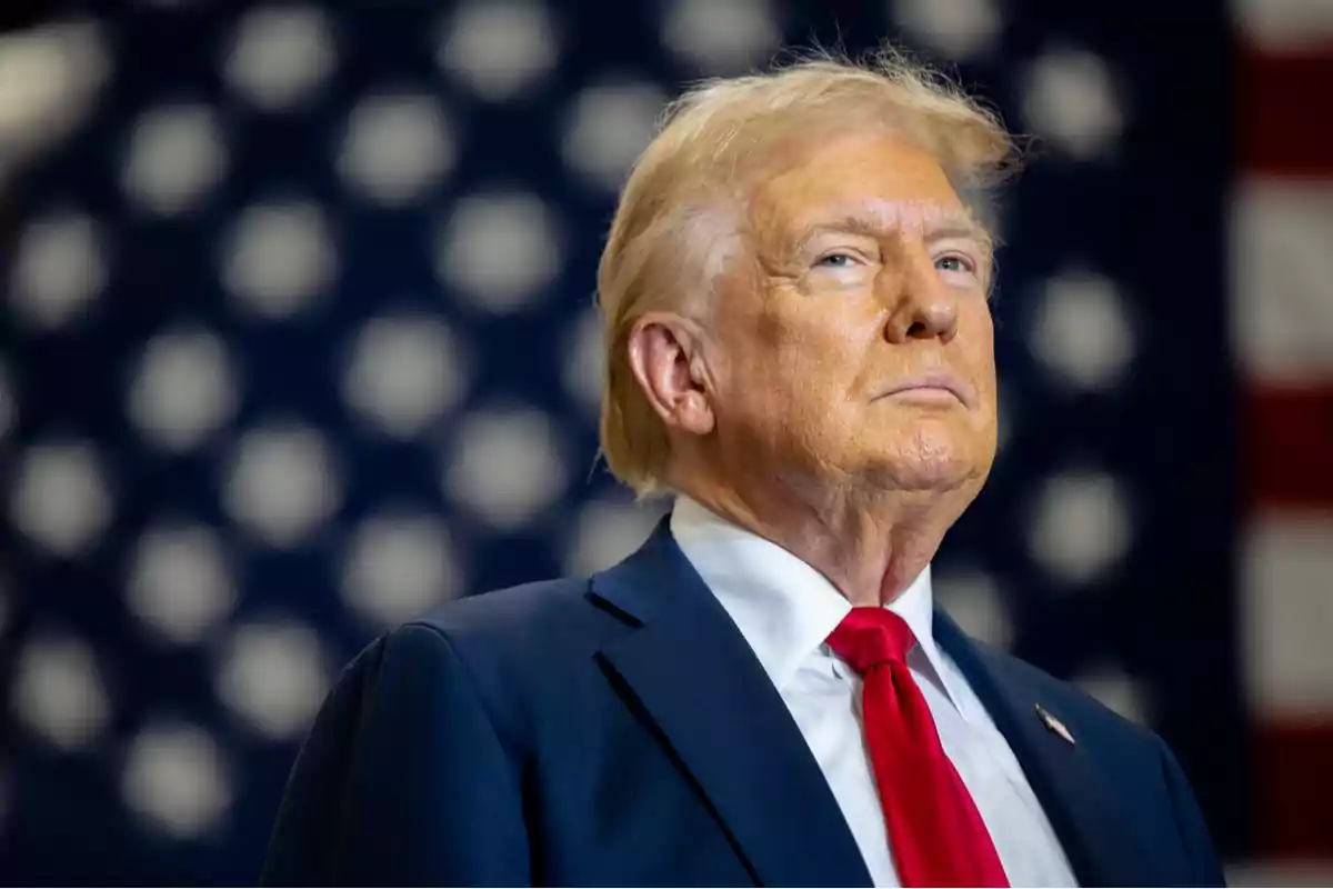 Blond-haired man in a blue suit and red tie in front of a blurred United States flag Blond-haired man in a blue suit and red tie in front of a blurred United States flag