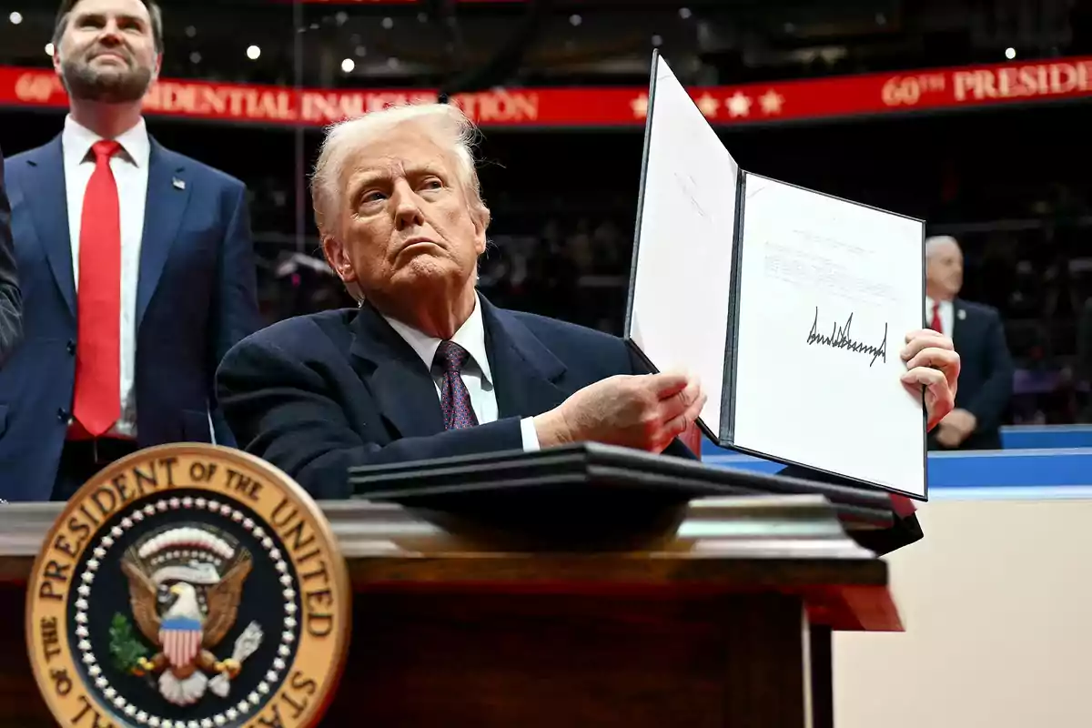 Un hombre de cabello rubio y traje oscuro muestra un documento abierto con una firma grande mientras está sentado en un escritorio con el sello presidencial de Estados Unidos al frente y varias personas de pie al fondo