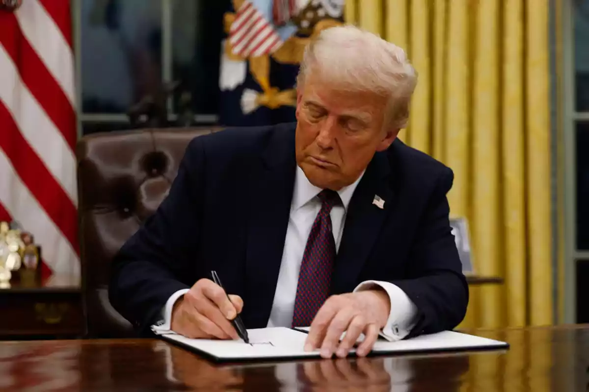 Blond-haired man in a dark suit signing a document at an office desk with yellow curtains and a U.S. flag in the background