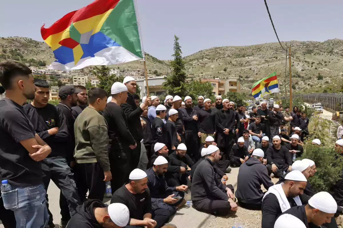 Grupo de personas reunidas al aire libre, muchas de ellas sentadas y usando gorros blancos, mientras ondean banderas de colores con un paisaje montañoso de fondo Grupo de personas reunidas al aire libre, muchas de ellas sentadas y usando gorros blancos, mientras ondean banderas de colores con un paisaje montañoso de fondo