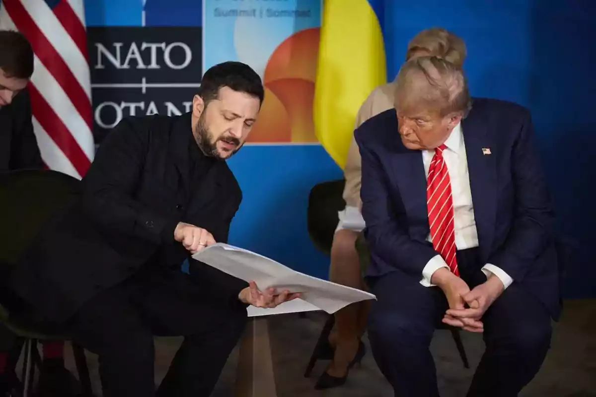Two men are seated and talking while one of them points to a document he is holding in his hands; in the background, flags and a NATO sign can be seen.