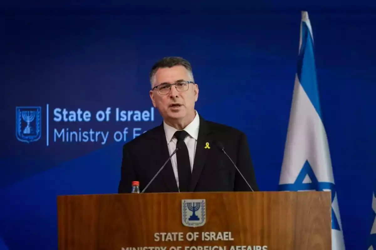 A man wearing glasses and a dark suit speaks at a podium of the Israeli Ministry of Foreign Affairs with an Israeli flag in the background.
