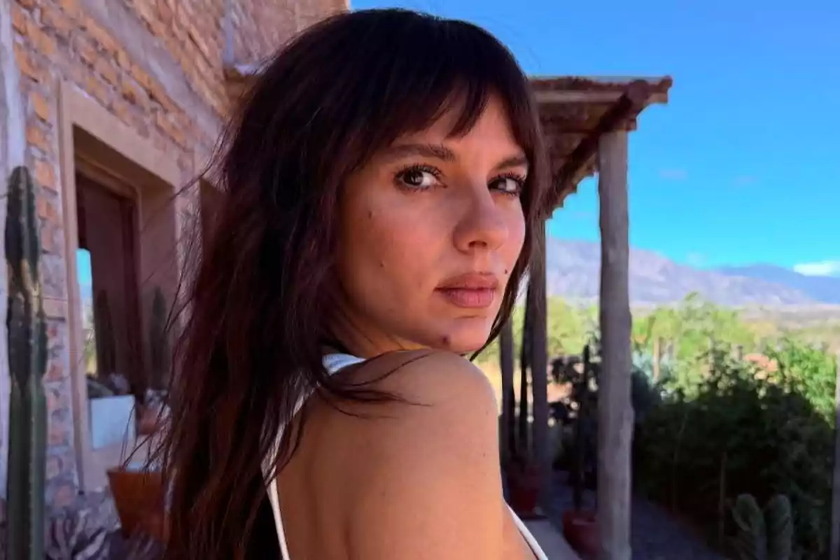 Mujer de cabello oscuro y largo mirando hacia la cámara con expresión seria al aire libre junto a una casa de ladrillo y vegetación de fondo