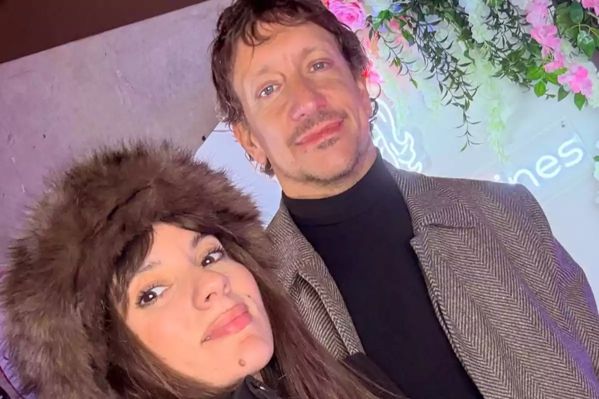 Una mujer con gorro de peluche y un hombre con saco posan juntos frente a una decoración de flores y luces.