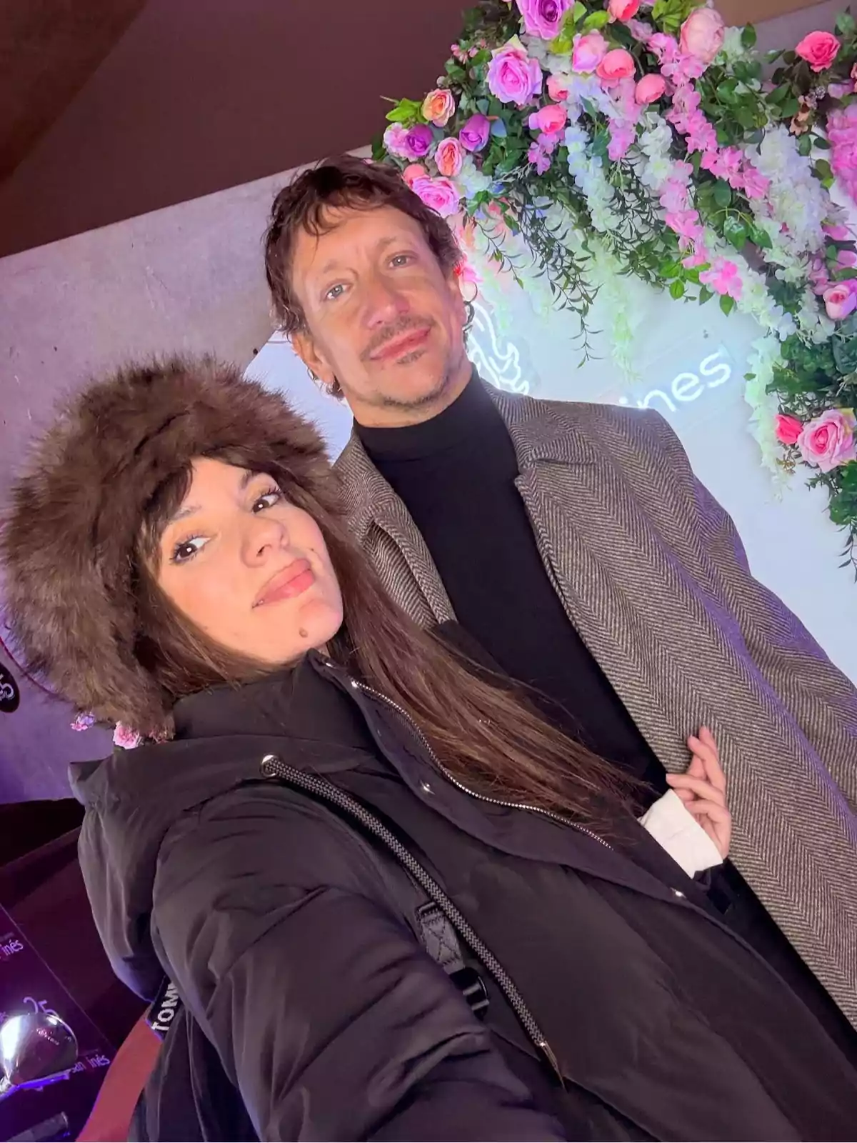 Two people pose for a selfie in front of a pink and white flower decoration, one wearing a black coat and a brown furry hat and the other a light gray coat. Two people pose for a selfie in front of a pink and white flower decoration, one wearing a black coat and a brown furry hat and the other a light gray coat.