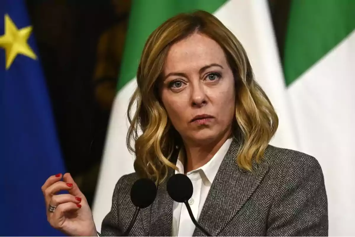 Mujer de cabello rubio y expresión seria hablando frente a micrófonos con banderas de fondo