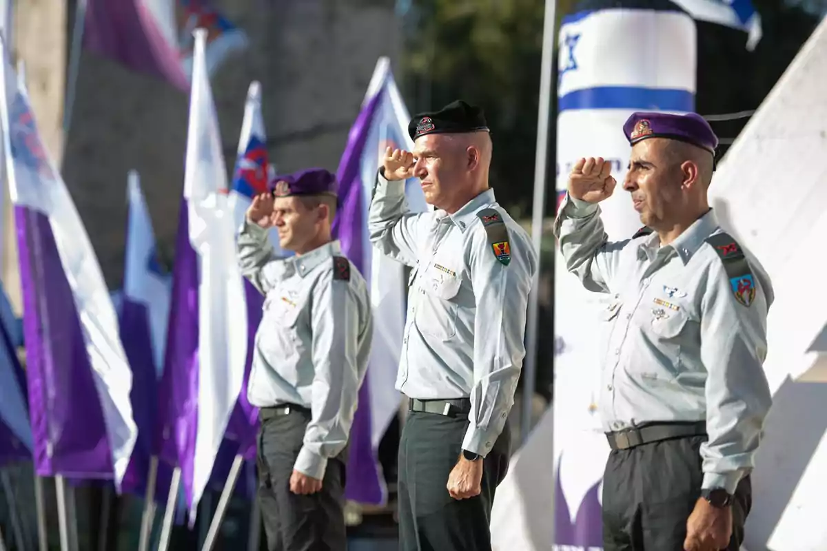 Tres militares uniformados hacen un saludo mientras están de pie en formación frente a varias banderas Tres militares uniformados hacen un saludo mientras están de pie en formación frente a varias banderas