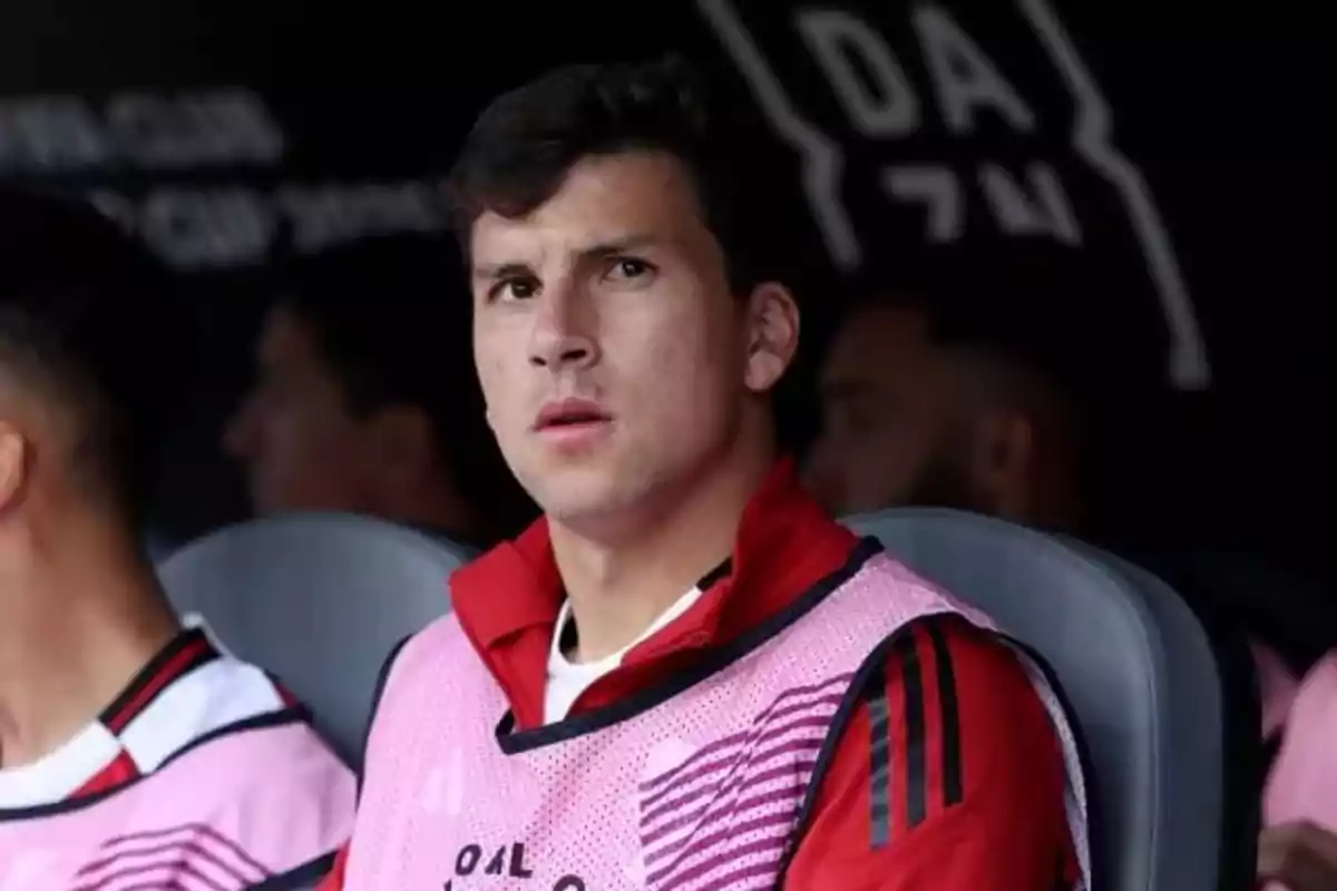 Young player sitting on the bench wearing a pink vest and a red jacket, looking straight ahead Young player sitting on the bench wearing a pink vest and a red jacket, looking straight ahead