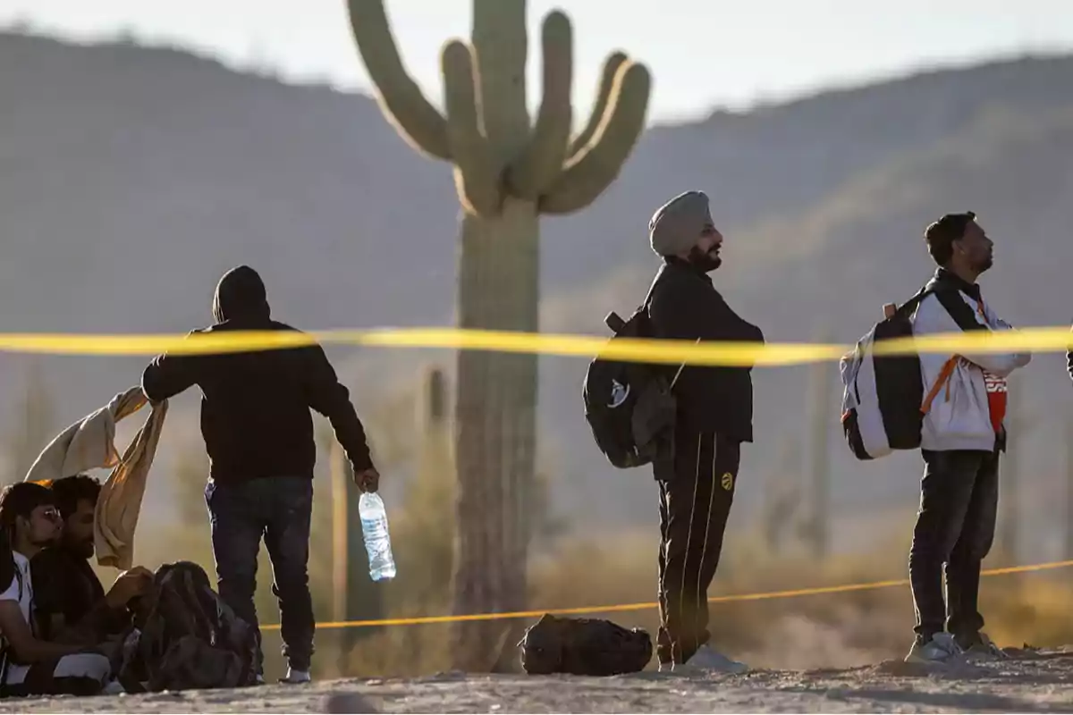 Varias personas con mochilas y botellas de agua esperan en el desierto junto a un cactus grande y una cinta amarilla en primer plano Varias personas con mochilas y botellas de agua esperan en el desierto junto a un cactus grande y una cinta amarilla en primer plano