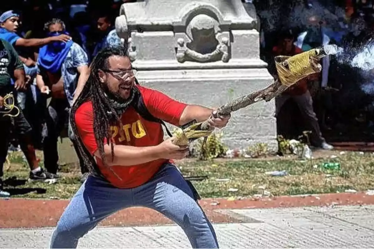 Hombre con camiseta roja y gafas sostiene un mortero artesanal mientras otras personas están al fondo en una manifestación al aire libre Hombre con camiseta roja y gafas sostiene un mortero artesanal mientras otras personas están al fondo en una manifestación al aire libre