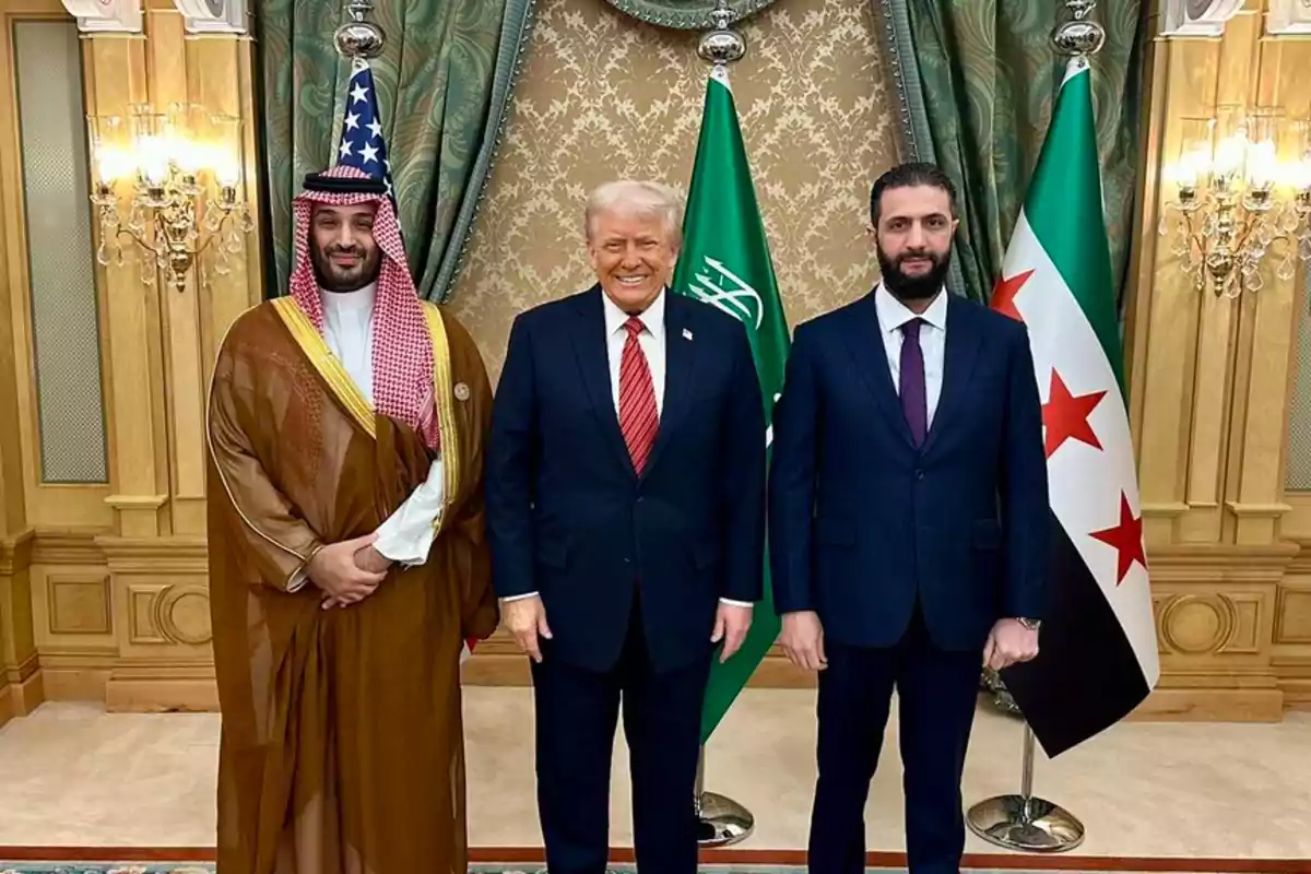 Three men stand posing for a formal photo in front of the flags of the United States, Saudi Arabia, and Syria in an elegantly decorated room. Three men stand posing for a formal photo in front of the flags of the United States, Saudi Arabia, and Syria in an elegantly decorated room.