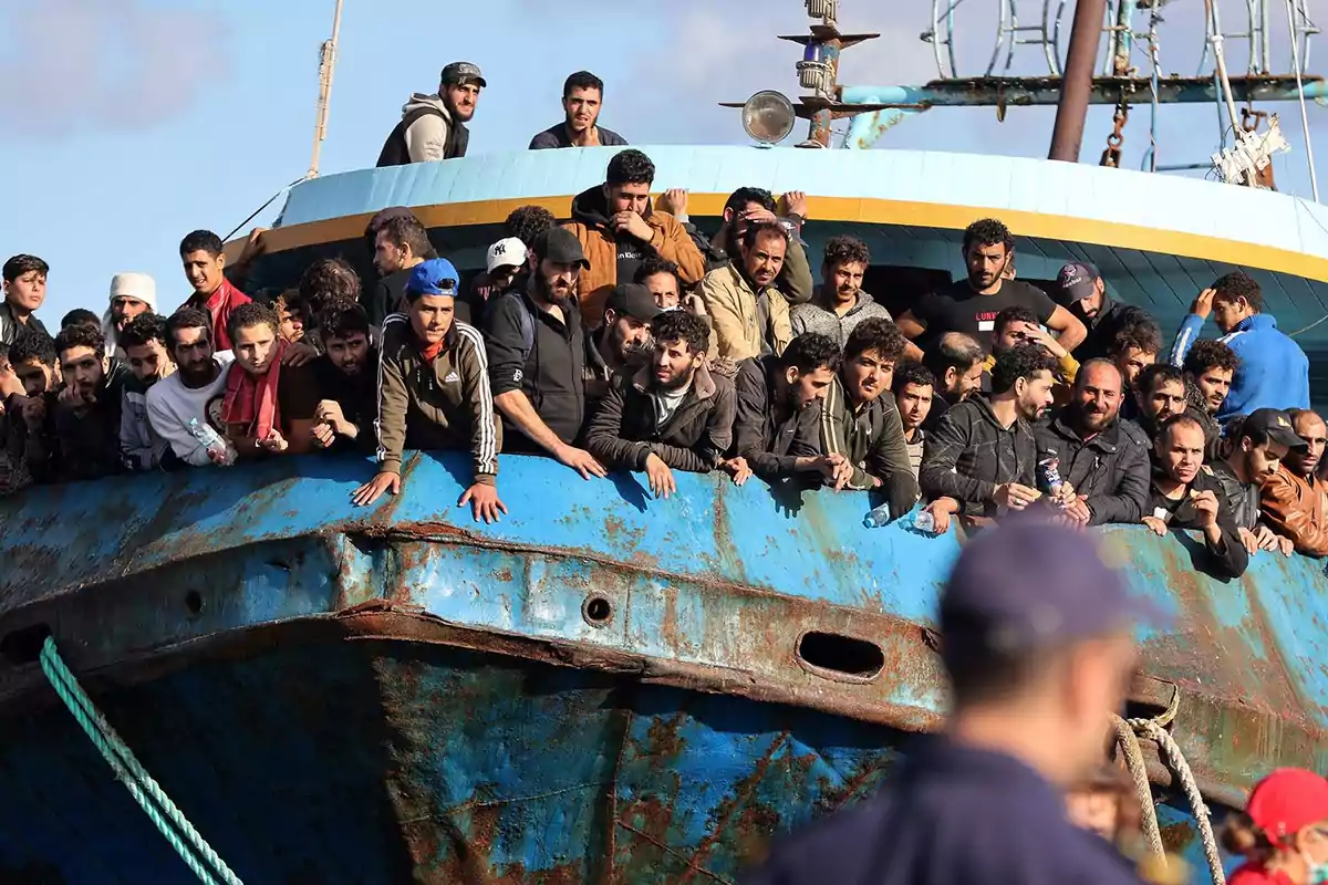 Un grupo numeroso de personas viaja en un barco oxidado y abarrotado, observando hacia la orilla mientras algunos oficiales se ven en primer plano.