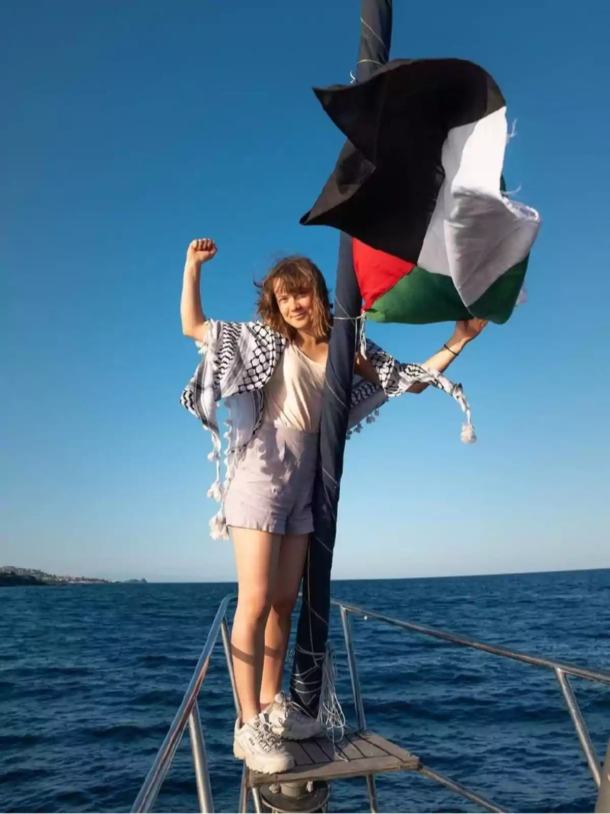 Mujer joven de pie en la proa de un barco levantando el puño junto a una bandera palestina ondeando al viento sobre el mar Mujer joven de pie en la proa de un barco levantando el puño junto a una bandera palestina ondeando al viento sobre el mar