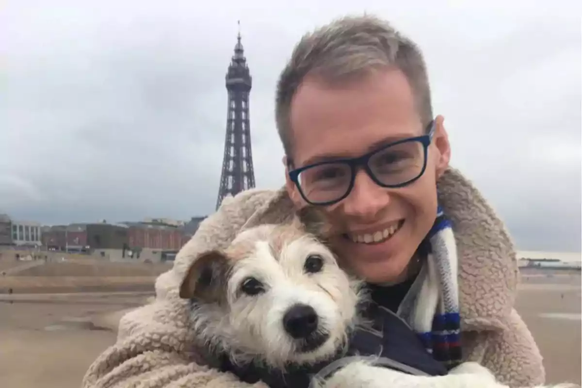 Hombre sonriente con gafas abrazando a un perro en la playa con una torre al fondo en un día nublado Hombre sonriente con gafas abrazando a un perro en la playa con una torre al fondo en un día nublado