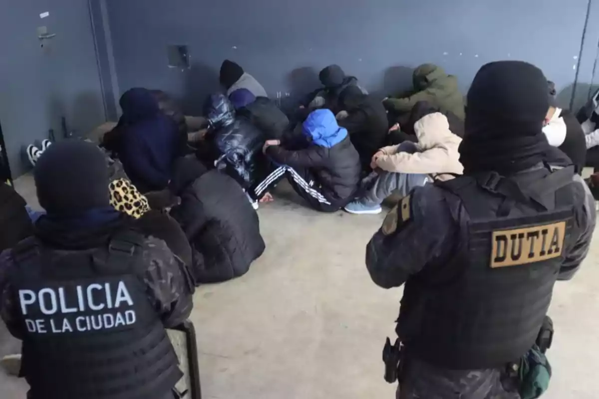 Group of people sitting on the floor with their heads down and two police officers watching over them in a closed room