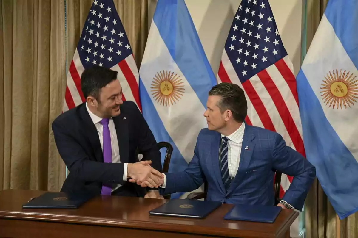 Two men in suits shake hands while seated at a table with folders in front of them and the flags of the United States and Argentina in the background. Two men in suits shake hands while seated at a table with folders in front of them and the flags of the United States and Argentina in the background.