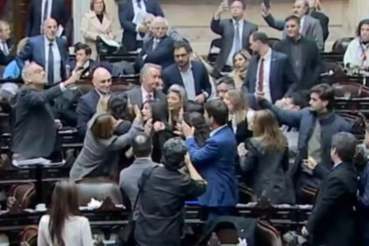 Un grupo de personas discute y se empuja en el recinto de un parlamento mientras otros observan y graban la escena con sus teléfonos móviles.