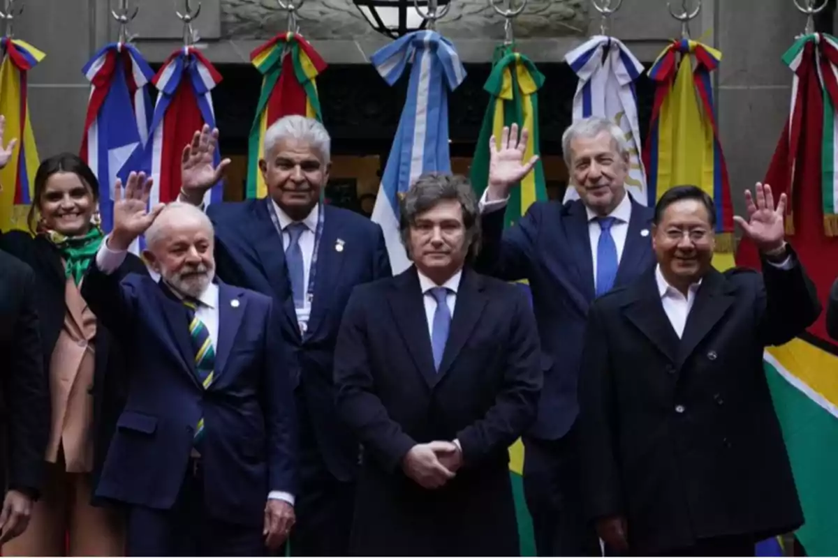 Grupo de personas vestidas formalmente posando frente a banderas de varios países sudamericanos mientras algunos de ellos saludan con la mano levantada