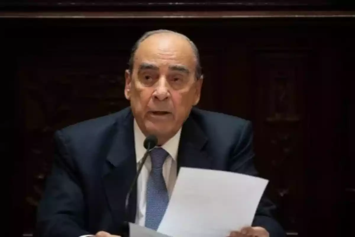 Older man in a dark suit speaking in front of a microphone and holding papers Older man in a dark suit speaking in front of a microphone and holding papers