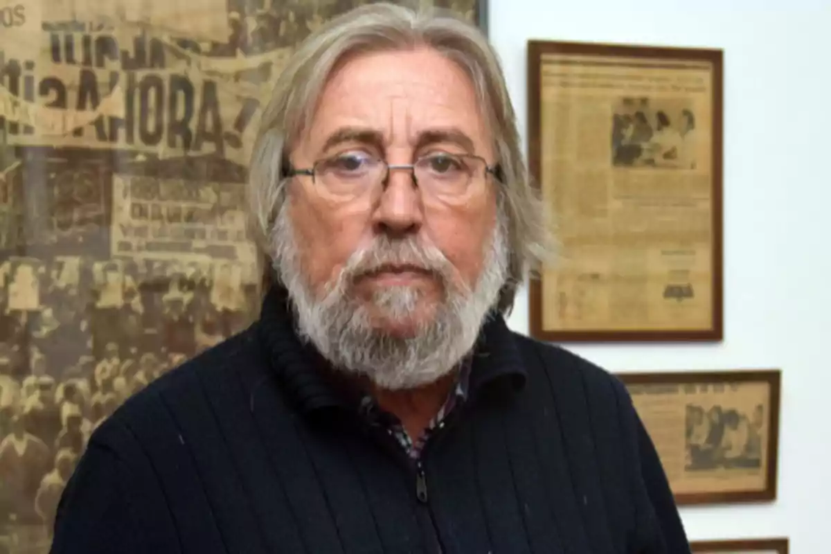 Gray-haired, bearded man with glasses and a dark sweater posing in front of a wall with framed newspaper clippings Gray-haired, bearded man with glasses and a dark sweater posing in front of a wall with framed newspaper clippings