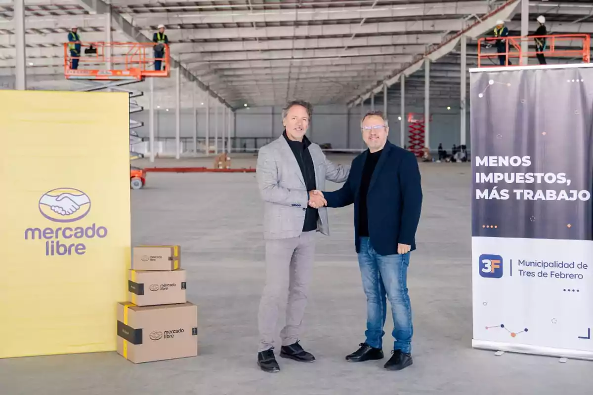 Dos hombres se dan la mano en el centro de un gran depósito en construcción, junto a cajas de Mercado Libre y banners promocionales Dos hombres se dan la mano en el centro de un gran depósito en construcción, junto a cajas de Mercado Libre y banners promocionales