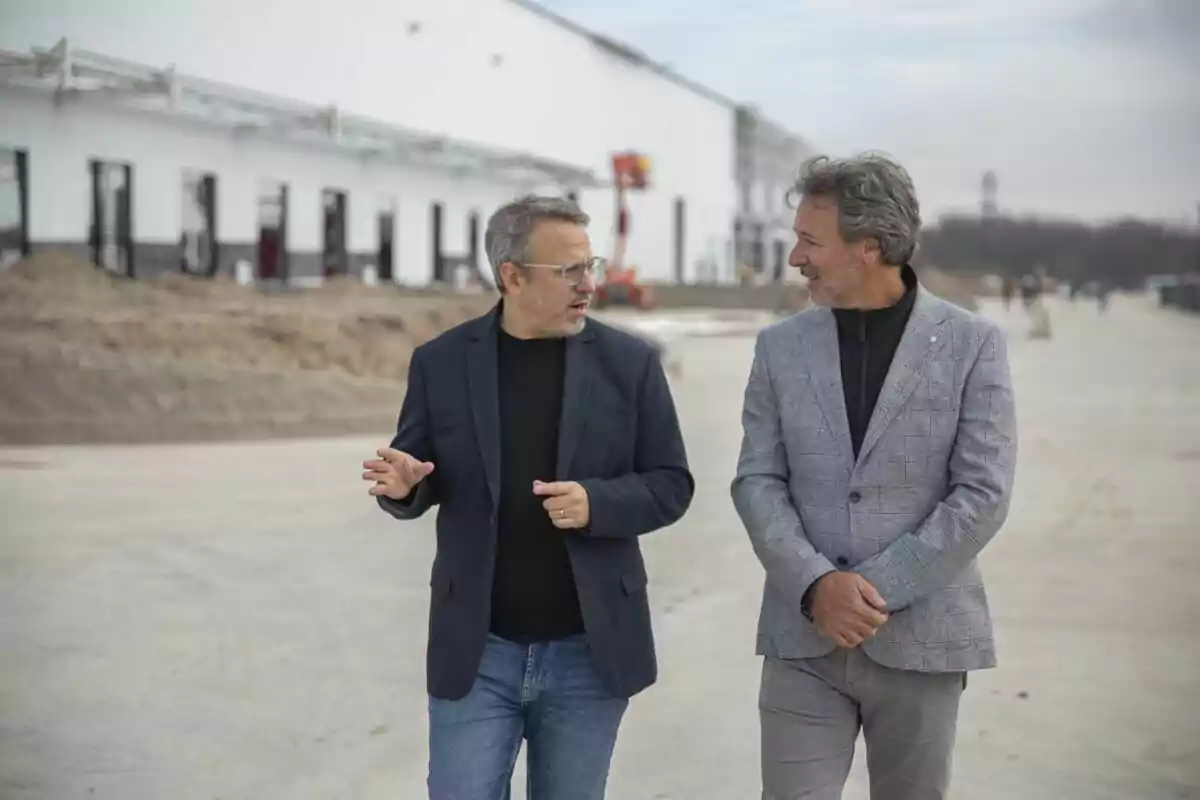 Dos hombres de mediana edad caminan y conversan frente a un edificio en construcción