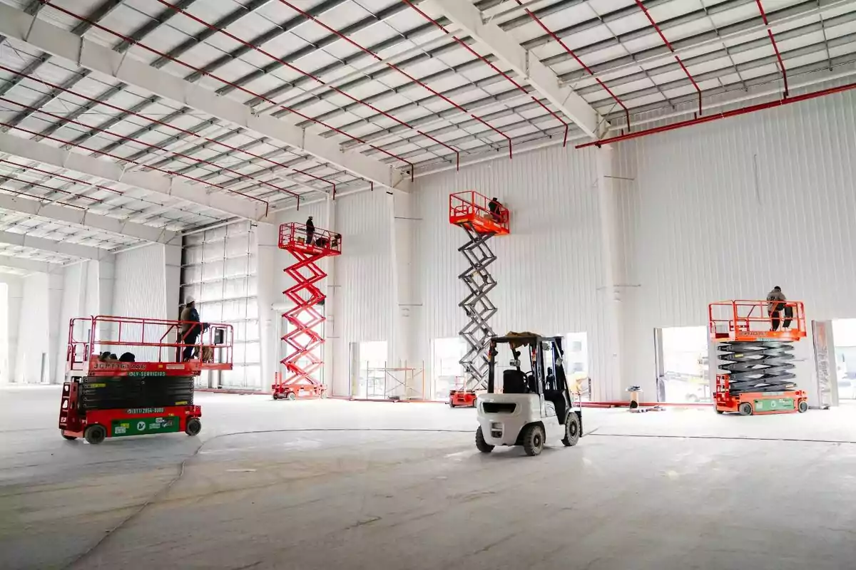 Trabajadores utilizando plataformas elevadoras y un montacargas dentro de una nave industrial en construcción Trabajadores utilizando plataformas elevadoras y un montacargas dentro de una nave industrial en construcción
