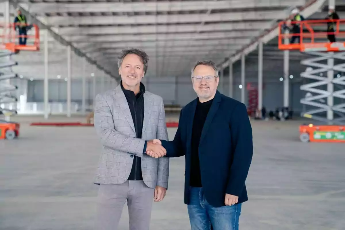 Dos hombres sonríen y se dan la mano en el interior de una nave industrial en construcción, con trabajadores y plataformas elevadoras al fondo.