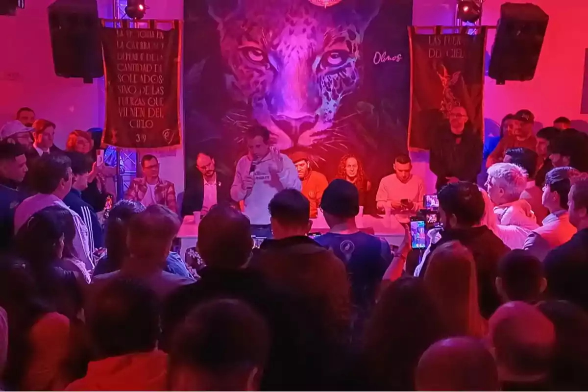A group of people gathered at an event with a mural of a jaguar in the background and colorful lights, while a man speaks into a microphone in front of a long table.