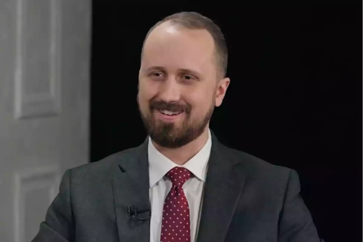 Hombre con barba y traje oscuro sonríe mientras está sentado frente a un fondo negro