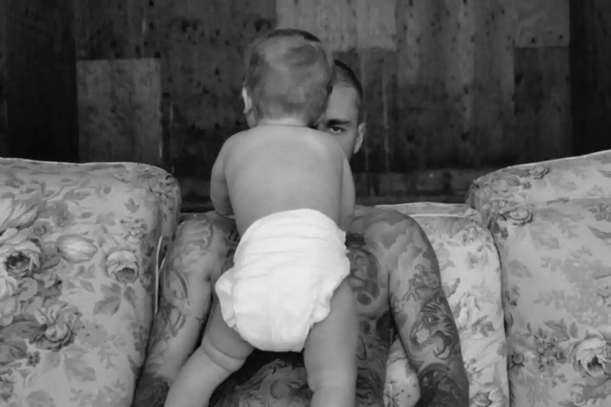 A tattooed man is sitting in a floral armchair while holding a diapered baby who has their back to the camera.