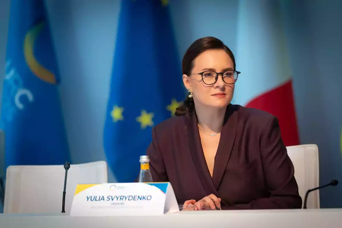 Mujer con gafas y saco oscuro sentada en una mesa de conferencia con banderas de fondo y un letrero con su nombre frente a ella Mujer con gafas y saco oscuro sentada en una mesa de conferencia con banderas de fondo y un letrero con su nombre frente a ella