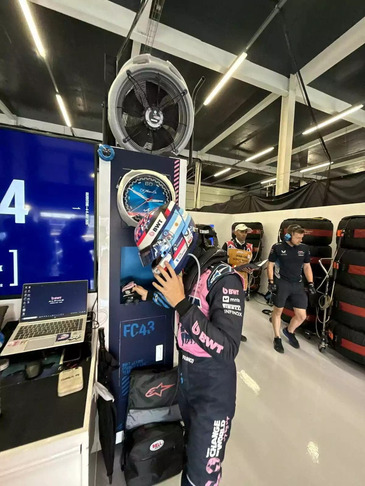 Auto racing driver with helmet and racing suit in a garage next to a large fan and technical equipment, while other people work in the background Auto racing driver with helmet and racing suit in a garage next to a large fan and technical equipment, while other people work in the background
