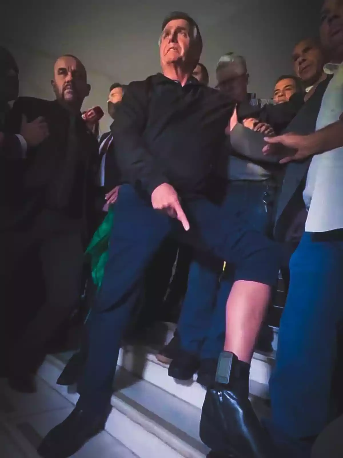 Un hombre rodeado de varias personas señala un brazalete electrónico en su tobillo mientras sube unas escaleras Un hombre rodeado de varias personas señala un brazalete electrónico en su tobillo mientras sube unas escaleras